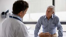 A patient sits on an exam table and listens to the doctor in the foreground talking A patient sits on an exam table and listens to the doctor in the foreground talking