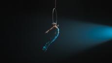 Trapeze artist in a blue costume holding onto a hoop in mid-air against a black backdrop Trapeze artist in a blue costume holding onto a hoop in mid-air against a black backdrop