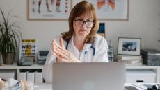A doctor with glasses sits at a desk and speaks during a telehealth visit. A doctor with glasses sits at a desk and speaks during a telehealth visit.